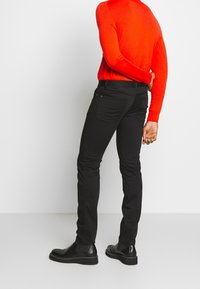 Persona che indossa un luminoso top rosso a maniche lunghe, pantaloni neri slim-fit e stivali in pelle neri, in piedi con una mano dietro la schiena.
