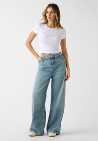 Biała krótka koszulka z logo w srebrnym stylu pisma, zestawiona z wysokim stanem, jasnoniebieskimi szerokimi spodniami i beżowymi tenisówkami. Gładka faktura, luźny krój.