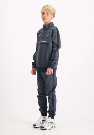 Navy Sportanzug aus glattem Stoff mit Reißverschlusskragen, Logo-Details und schwarzen Seitenstreifen; kombiniert mit weißen Sneakern mit schwarzen Akzenten.