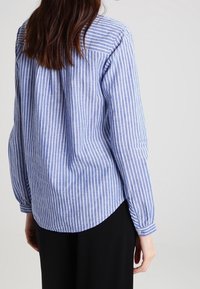 Chemise rayée bleue et blanche à manches longues, avec des poignets boutonnés, un empiècement au dos et un ourlet arrondi. Le tissu semble léger avec une texture lisse.