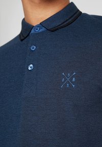 Marinblå polo med texturerad tyg, en kontrasterande svart krage, tre blå knappar och en ljusblå broderad logotyp på bröstet.