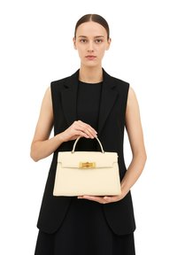 Crème leren handtas met een gestructureerd ontwerp, gouden hardware en een bovenhandvat. Gepresenteerd door een model in een zwart outfit.
