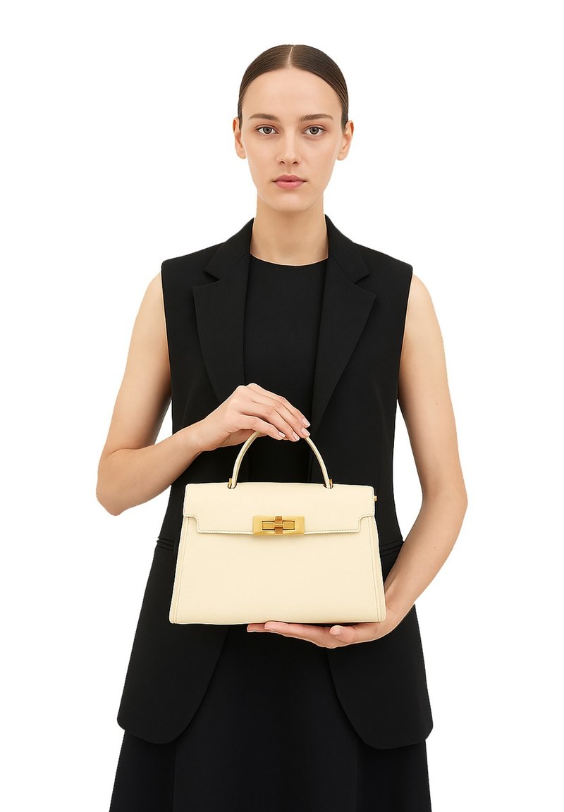 Crème leren handtas met een gestructureerd ontwerp, gouden hardware en een bovenhandvat. Gepresenteerd door een model in een zwart outfit.