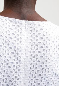 Biała sukienka z otwartym wzorem haftu, wyposażona w zamek błyskawiczny na środku pleców, podkreślająca teksturowaną tkaninę z owalnymi wycięciami.