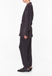 Chaqueta larga de ante negra con cinturón, combinada con pantalones holgados negros y mules negros de tacón, presenta una textura sencilla y suave.