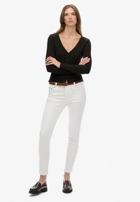 Mujer de pie con los brazos cruzados, vistiendo una blusa negra de manga larga con botones, pantalones ajustados blancos, cinturón marrón y mocasines marrones oscuros sobre fondo blanco.