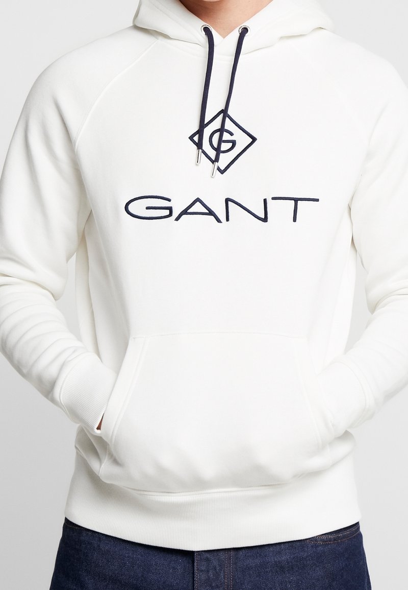 Sudadera blanca con bolsillo canguro, puños y dobladillo acanalados. Presenta un logo de GANT en azul marino y un diseño de diamante en el pecho. Material de mezcla de algodón.