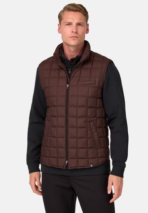 Chaleco acolchado marrón con cuello alto, cremallera frontal, bolsillos laterales y un diseño ligero, superpuesto a una camiseta de manga larga negra.