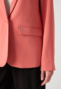 Korallfärgad blazer i texturerat tyg med en enkel bröstficka med detaljer i silverknapp och en skräddarsydd passform, matchad med svarta byxor.