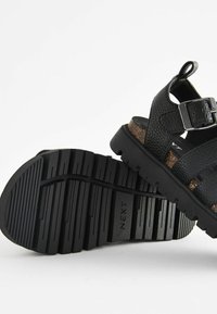 Svarta sandaler med en texturerad ovandel, justerbar spänne och korkfotbädd. Skulpterad svart gummisula för grepp och hållbarhet.