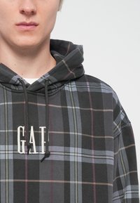 Sudadera con capucha en un patrón de cuadros negro y gris. Presenta texto bordado en blanco "GAF" en el pecho y cordones en la capucha.