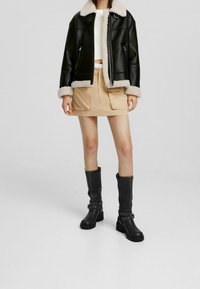 Kvinna iklädd svart shearlingjacka, vit kortärmad tröja, beige kjol med fickor och svarta stövlar som når mitt på vaden med remmar, stående mot grå bakgrund.