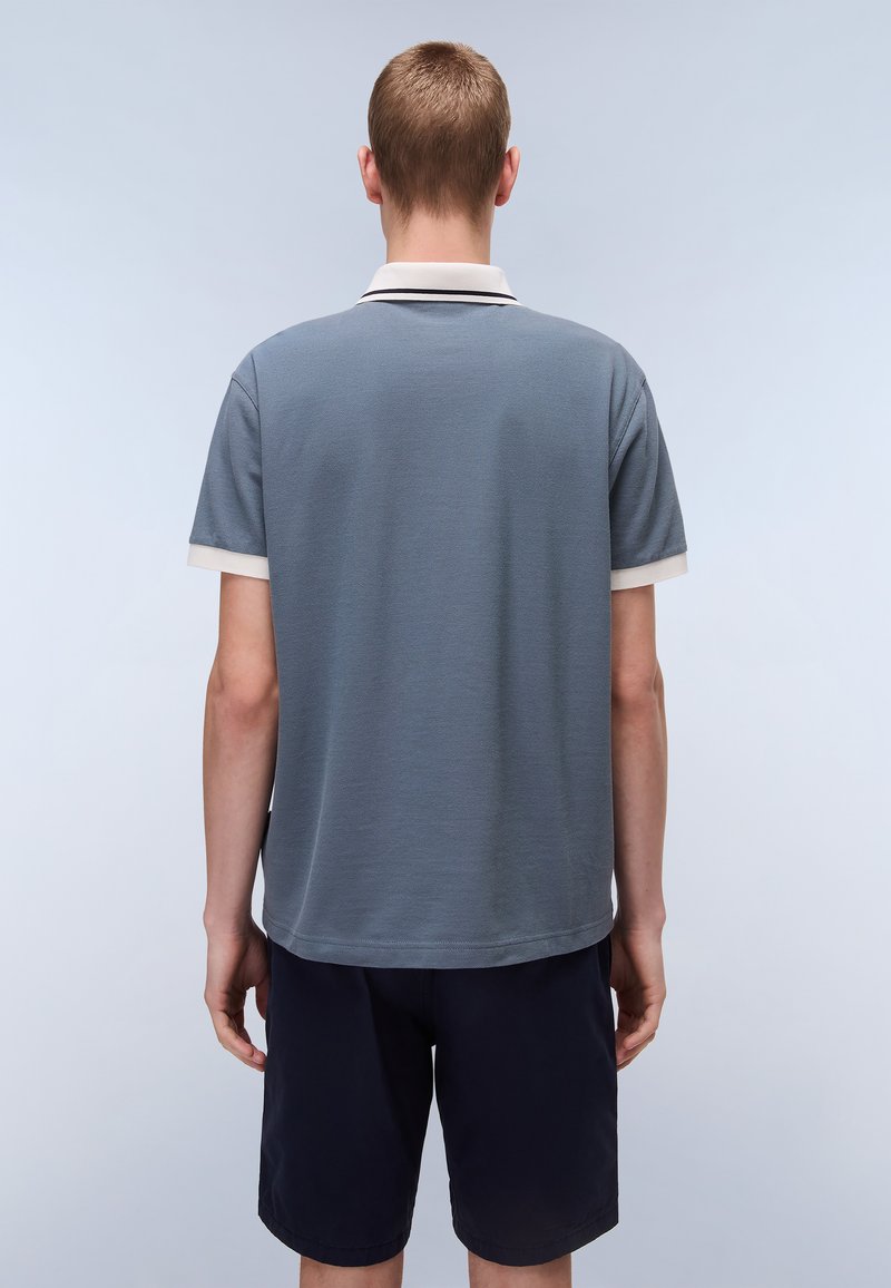 Polohemd in Schieferblau mit strukturierter Oberfläche, mit einem weißen Kragen und Ärmelausschnitten. Kombiniert mit dunklen Marineblauen Shorts.