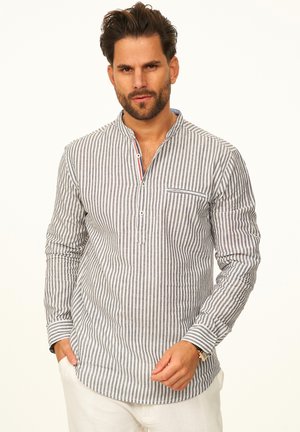 Mann trägt ein langärmliges Hemd mit vertikalen Streifen und Mandarin-Kragen sowie eine weiße Hose, steht mit einer Hand in der Tasche.