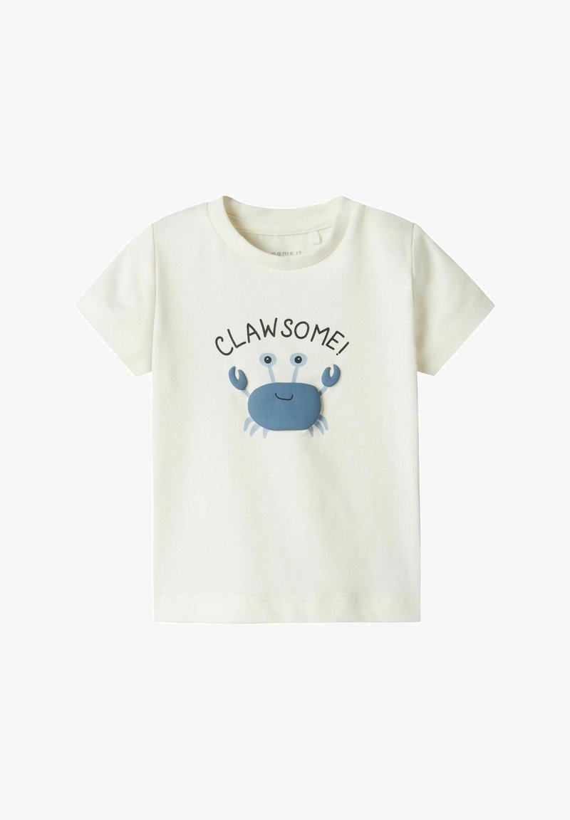 Camiseta de algodón color crema claro con un gráfico de un cangrejo azul y la palabra "¡GARRA-SÓLIDO!" impresa arriba en negro. Diseño de manga corta y cuello redondo.