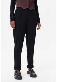 Zwarte taps toelopende broek met plooien, voorzien van zijzakken. Gecombineerd met bruine loafers, wordt de look afgemaakt met een strakke bruine gilet.