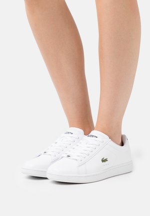 Personne portant des baskets blanches Lacoste à lacets, avec un logo crocodile vert et des semelles blanches sur un fond uni.