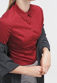 Chemise rouge boutonnée avec col, texture lisse et coupe ajustée, associée à un vêtement d'extérieur gris fluide.