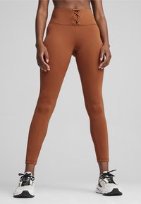 Højtaljede brune leggings med snørelukning foran, lavet af strækbart materiale, parret med sort-hvide sneakers.