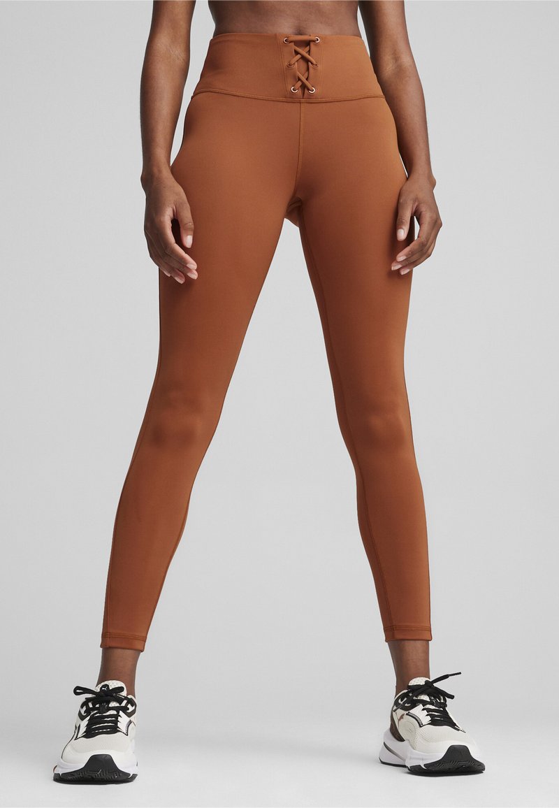 Højtaljede brune leggings med snørelukning foran, lavet af strækbart materiale, parret med sort-hvide sneakers.