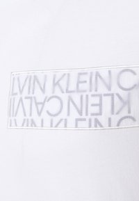 Valkoinen puuvillapaita, jossa on suorakulmainen logopatchi, jossa toistuva teksti "CALVIN KLEIN" vaaleanharmaana läpinäkyvällä taustalla.