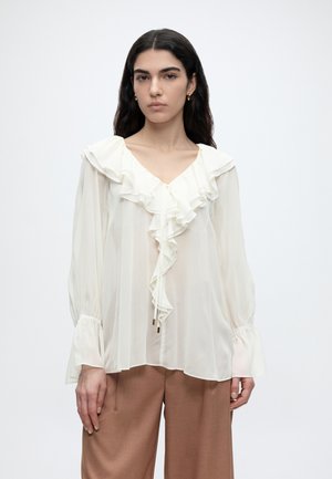 Kobieta o długich ciemnych włosach nosi białą przezroczystą bluzkę z falbankami przy dekolcie w serek i długimi rękawami, połączoną z jasnobrązowymi szerokimi spodniami na białym tle.