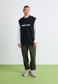 Sort ærmeløs sweatshirt med "NIKE AIR" grafik, båret over en hvid langærmet trøje. Olivengrønne cargo-bukser og grå sportsko fuldender looket.