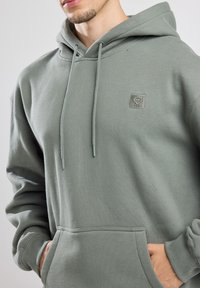 Haut à capuche vert clair en matériau doux, avec une poche avant, des cordons de serrage et un petit logo brodé sur la poitrine.