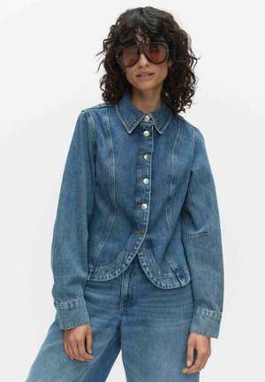 Kvinna med lockigt hår som bär stora solglasögon, en blå jeansjacka med knappar och matchande jeans mot en enfärgad bakgrund.