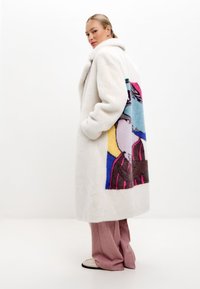 Vit fuskpälsrock med en stor färgglad grafisk design på baksidan. Har en sjalkrage och sidofickor. Skräddarsydd passform och knälång.