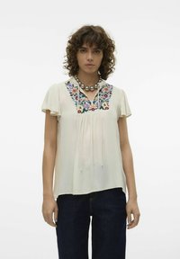 Lys beige bluse med blomsterbroderi i pink, blå og grøn ved halsudskæringen. Korte ærmer og afslappet pasform. Bæres med mørkeblå jeans.