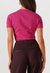 Aansluitend roze t-shirt met korte mouwen en oranje stiksels. Gepaard met donkerbruine broek met achterzakken en een logo-detail.