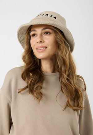 Vrouw met lang golvend haar die een beige bucket hat en bijpassende beige trui draagt, licht glimlachend en zijwaarts kijkend.