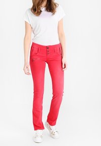 Donna con pantaloni aderenti rossi brillanti dotati di più bottoni e tasca con zip, t-shirt bianca e sneakers bianche, in piedi.