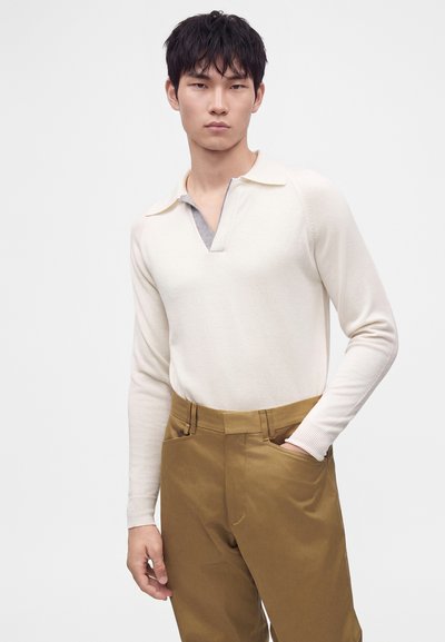 Jeune homme portant un polo à manches longues crème et un pantalon marron, une main dans la poche, sur un fond clair uni.