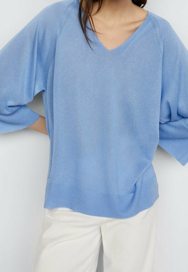 Femme portant un pull ample bleu clair à col en V avec de larges manches et un pantalon blanc cassé, debout les mains sur les hanches.