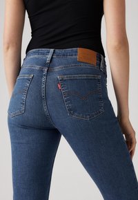 Tumesinised teksapüksid keskmise vöökohaga, millel on tagataskud, oranž õmblus ja pruun nahast plaastriga vöörihm.