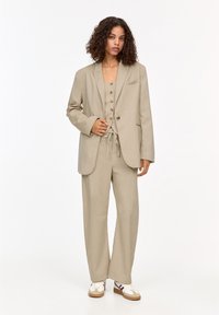 Beige pinstriped suit with a tailored blazer, matching vest with buttons, and wide-leg pants, complemented by white sneakers.