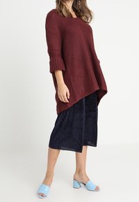 Burgundy stickad tröja med sänkta axlar, ihop med en marinblå plisserad kjol och ljusblå klacktofflor med blockklack. Texturerade material ingår.