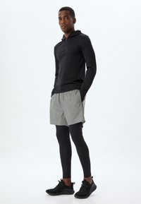 Sort langærmet zip hoodie, lysegrå shorts over sorte leggings, og sorte sportsko. Glat stof, tætsiddende design og afslappet stil.