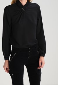 Svart, långärmad blus med hög krage och en liten silverdetalj vid halsringningen. Bärs med svarta cargobyxor med dragkedjor.