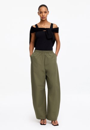 Femme portant un haut noir sans manches et un pull noué sur les épaules, avec un pantalon ample vert olive et des talons ouverts noirs, debout les mains dans les poches.