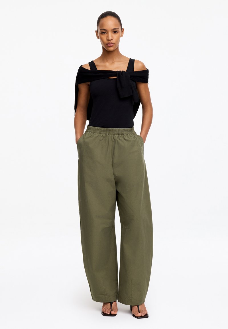 Femme portant un haut noir sans manches et un pull noué sur les épaules, avec un pantalon ample vert olive et des talons ouverts noirs, debout les mains dans les poches.