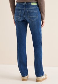 Jeans en denim bleu avec jambes droites, cinq poches, délavage usé et une étiquette de marque verte sur la taille arrière. Tissu texturé.