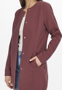 Lång vinröd kappa med rund halsringning, en rad silverfärgade tryckknappar, två sidfickor och ett slätt, texturerat tyg.