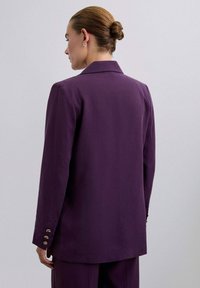 Blazer púrpura con una forma estructurada y cuello con muescas. Presenta detalles de botones dorados en las mangas, confeccionado en una tela suave con un acabado mate.