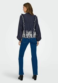 Tumed blouse volangide accentidega, valge lilleline pitsääris, koos siniste denim-pükstega ja mustade plokknööpadega kingadega.
