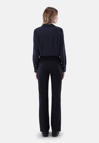Frau mit blonden Haaren, die ein dunkelblaues, gepunktetes Langarmshirt und schwarze Hosen trägt, steht mit dem Rücken zu einer weißen Wand.