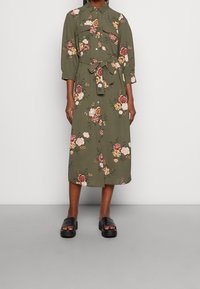Olivgrünes Midikleid mit Blumenmuster, Kragen, Knopfleiste, verstellbarem Taillengürtel und Dreiviertelärmeln. Kombiniert mit schwarzen Sandalen.