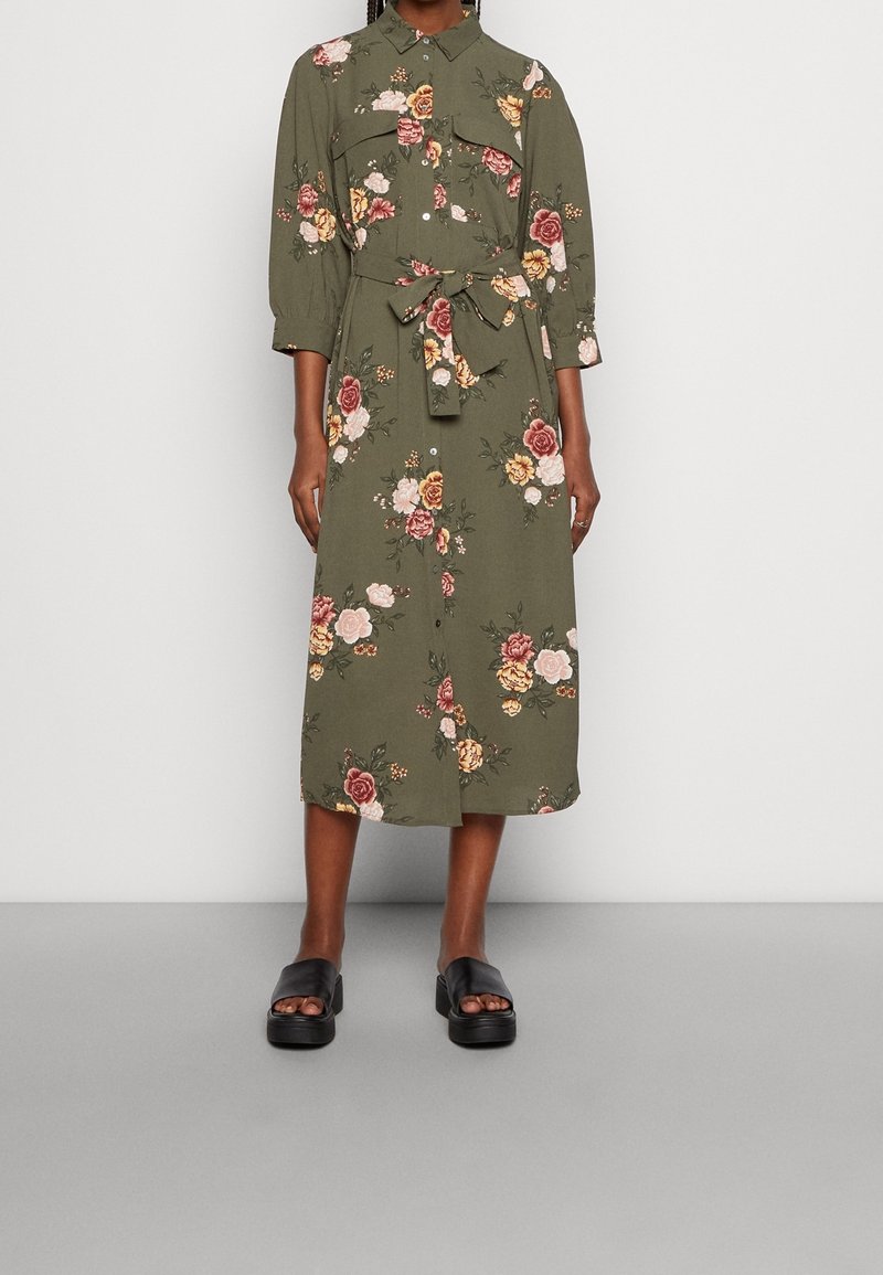 Olivgrünes Midikleid mit Blumenmuster, Kragen, Knopfleiste, verstellbarem Taillengürtel und Dreiviertelärmeln. Kombiniert mit schwarzen Sandalen.